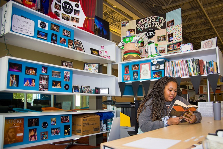 Ophelia Griffin sits in a chair on the right side of the image, reading a book. She's in a large nook that has shelves on two sides of her, filled with displays and props for the student theatre group.